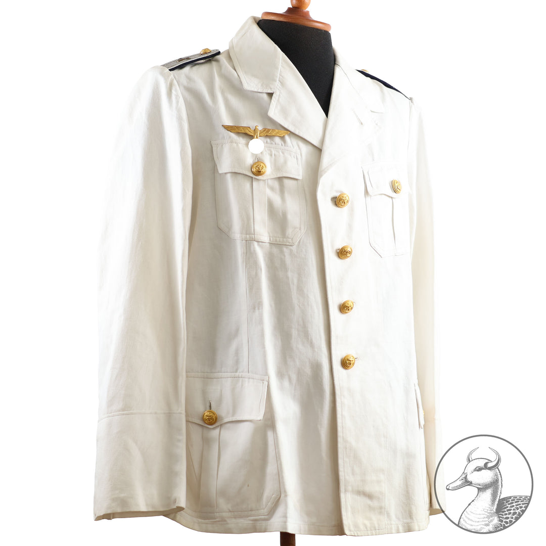 Weißes Jackett (nach neuer Vorschrift) der Kriegsmarine für einen Leutnant der
Küstenartillerie . Das Jackett besitzt aufgesetzte Brust- und Seitentaschen mit Quetschfalten. Die
feuervergoldeten Ankerknöpfe sind sicher mit Sprengringen angebracht, während der
vorschriftsmäßige Metallbrustadler mittels typischer Zwirnschlaufen befestigt ist. Innen finden sich ein Größen- und Herstellerstempel sowie die Namensstickerei
„Hadenfeld“
