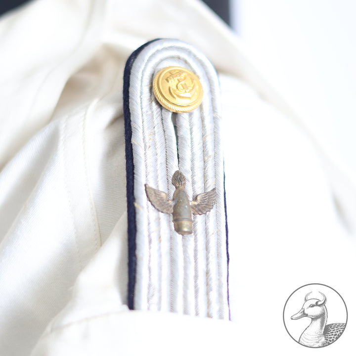 Weißes Jackett (nach neuer Vorschrift) der Kriegsmarine für einen Leutnant der
Küstenartillerie . Das Jackett besitzt aufgesetzte Brust- und Seitentaschen mit Quetschfalten. Die
feuervergoldeten Ankerknöpfe sind sicher mit Sprengringen angebracht, während der
vorschriftsmäßige Metallbrustadler mittels typischer Zwirnschlaufen befestigt ist. Innen finden sich ein Größen- und Herstellerstempel sowie die Namensstickerei
„Hadenfeld“